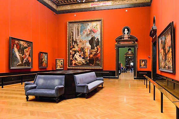 Roter barocker Museumssaal mit Couches in der Mitte und Gemälden an der Wand