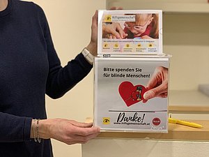 von Person gehaltene Spendenbox mit Aufschrift: Bitte spenden Sie für blinde Menschen. Danke! www.hilfsgemeinschaft.at. rotes Herz mit Maulwurf-Cartoon und Langstock
