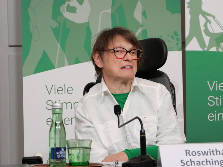 Eine Frau mit braunem kurzen Haar und weißer Jacke sitzt auf einem Podium mit grünem Banner im Hintergrund.