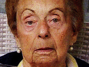 Eine ältere Dame mit Perlenohrringen und hellem Tuch um den Hals blickt gerade in die Kamera