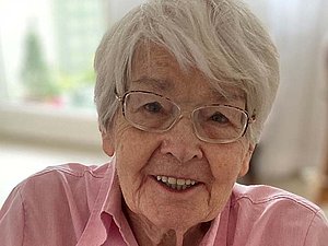 Porträt von Dame mit weißen Haaren und Brille in rosa Bluse, die in die Kamera blickt.