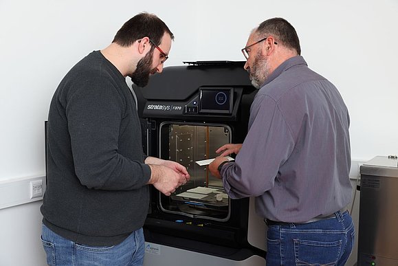 zwei Männer stehen vor 3D Drucker und sind über einen weißen Gegenstand mit Bebraillung gebeugt
