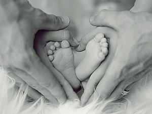 Vier Hände bilden ein Herz um die Füße eines Babys.