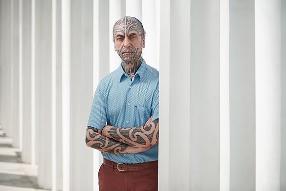 Mann mit Gesichtstattooos im blauen Hemd schaut hinter weißen Säulen hervor