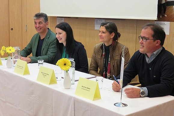 Podium mit zwei Frauen und zwei Männern, die bei einem langen Tisch sitzen. 