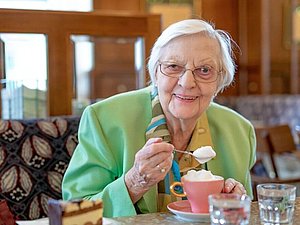 Frau J. sitzt einem Café, in der Hand einen Löffel mit Milchschaum, vor sich am Tisch eine Melange sowie ein Stück Kuchen.