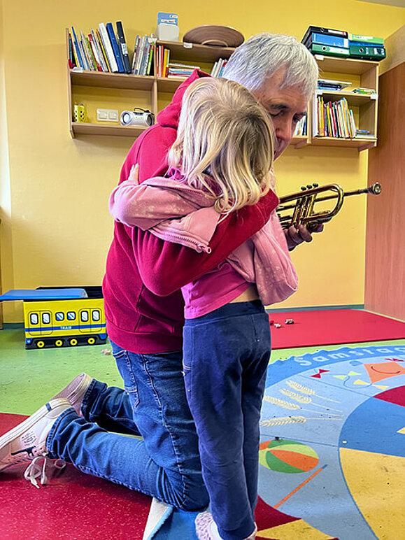 Älterer Mann und kleines Mädchen umarmen sich in einem Spielzimmer, er kniet und hält ein Blasinstrument in der Hand.