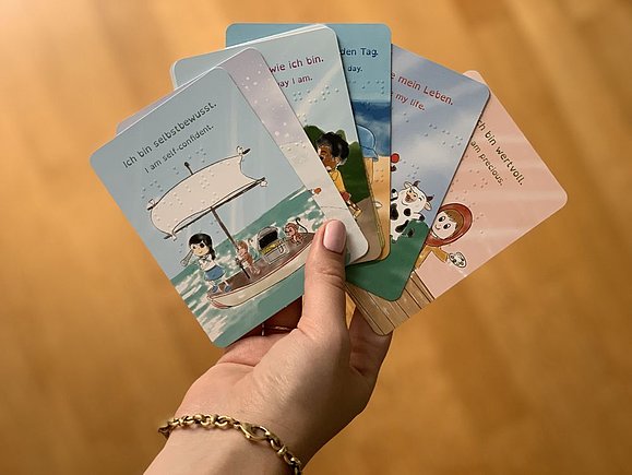 Hand hält mehrere Karten in der Hand mit Illustrationen von Kindern und Tieren mit Sprüchen wie: Ich bin selbstbewusst (Mädchen als Piratin am Schiff).