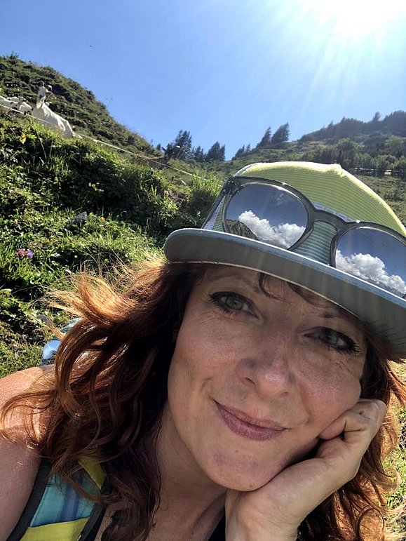 Selfie einer jungen Frau mit Basecap und Sonnenbrille auf, im Hintergrund grüner Berghang und Gams