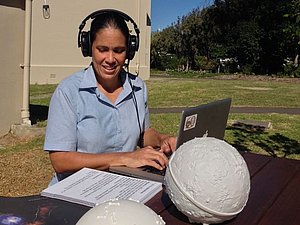 Eine Frau sitzt bei einem Tisch vor einem Planetarium. Sie bedient einen Laptop und hat dabei Kopfhörer auf. Daneben liegen 3D Abbildungen von Himmelskörpern und ihre Notizen.