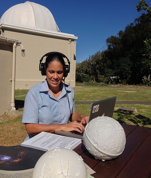 Eine Frau sitzt bei einem Tisch vor einem Planetarium. Sie bedient einen Laptop und hat dabei Kopfhörer auf. Daneben liegen 3D Abbildungen von Himmelskörpern und ihre Notizen.