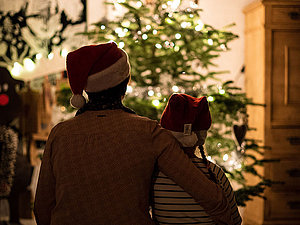 Ein großer und ein kleiner Mensch mit Weihnachtsmütze sitzen vor einem hellerleuchteten Weihnachtsbaum.