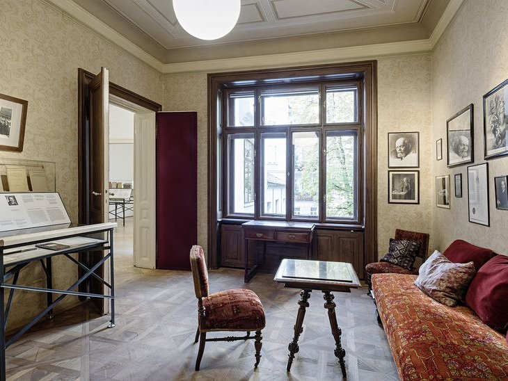 Ein Zimmer mit großen Fenstern, Bildern an der Wand, einem alten Schreibtisch direkt beim Fenster, einer Couch, einem Tisch und 2 Polsterstühlen. Links führt eine Tür ins nächste Zimmer.
