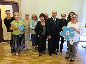 Gruppenfoto von 10 Personen in einem Raum, 2 Damen halten blaue Vergissmeinnicht-Blumen aus Papier in den Händen. 