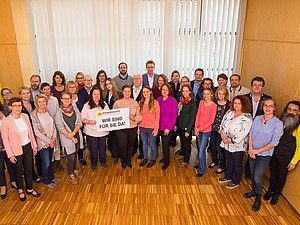 Ungefähr 40 Menschen stehen im Halbkreis nebeneinander. in einem Raum Ganz vorne stehen zwei Frauen mit einem Schild mit der Aufschrift "Wir sind für Sie da".