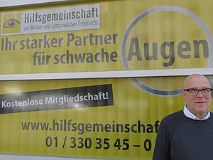 Mitarbeiter Daniele M. steht vor einem gelbschwarzen Plakat der Hilfsgemeinschaft