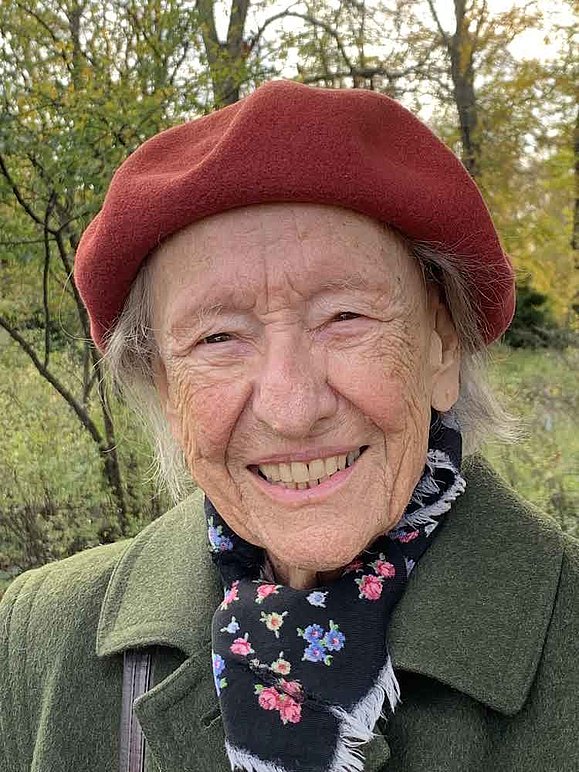 ältere Dame mit rostroter Haube und grüner Filzjacke vor Wald lächelt in die Kamera