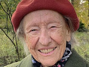ältere Dame mit rostroter Haube und grüner Filzjacke vor Wald lächelt in die Kamera