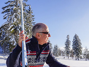 Ein älterer Mann mit Norvegerpulli und Sonnenbrille sitzt in einer Winterlandschaft und schaut lächelnd in den Himmel