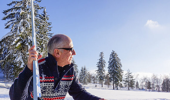 Ein älterer Mann mit Norvegerpulli und Sonnenbrille sitzt in einer Winterlandschaft und schaut lächelnd in den Himmel