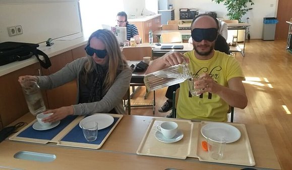 Eine Frau und ein Mann mit Augenbinden versuchen Glas und Tasse mit Wasser zu füllen