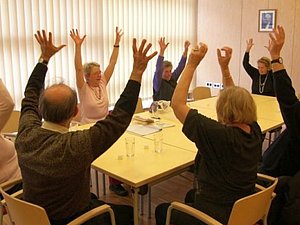 Mehrere Menschen sitzen rund um einen großen Tisch. Alle strecken die Arme in die Höhe.