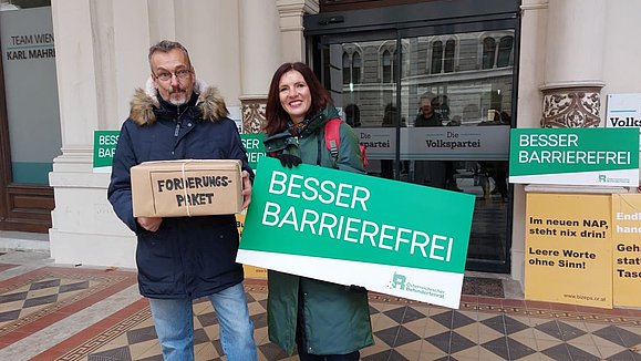 Mann mit Kiste "Forderungspaket" und Frau mit grünem Schild "Besser barrierefrei" vor Hauseingang
