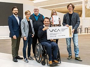 Gruppe von Menschen mit Anzug lächeln in die Kamera. Einer sitzt im Rollstuhl und hält einen überdimensionalen Scheck mit Aufschrift: WACA Zertifikat - Bronze