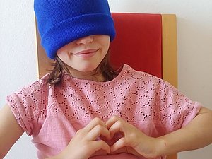 Ein Mädchen mit einer blauen Mütze auf dem Kopf und vor den Augen formt mit ihren Händen ein Herz
