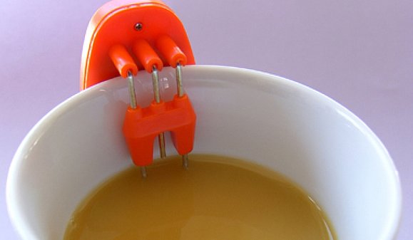 Tasse mit Kaffe und rotem Gerät geklammert an den Tassenrand, reicht bis in die Flüssigkeit hinein.