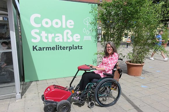 Maria Grundner sitzt im Rollstuhl vor der grünen Plakatwand der "Coolen Straße" am Karmeliterplatz.