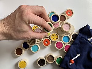 Hand hält Holzring mit blauem Stoff innen in der Hand und berührt Stoff mit dem Finger, dahinter ein ausgeleerter blauer Beutel und viele bunte Holzringe