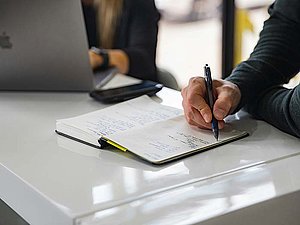 Eine Männerhand macht Notizen in einem aufgeschlagenen Notizbuch auf einem Tisch, im Hintergrund sieht man teilweise eine Frau, die an einem Laptop sitzt.