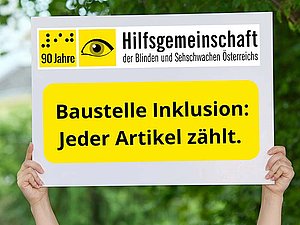 Zwei Hände halten ein weißes Schild vor grünem, unscharfem Hintergrund (Bäume und Natur). Auf dem Schild oben links das Logo der „Hilfsgemeinschaft der Blinden und Sehschwachen Österreichs“ mit der Aufschrift „90 Jahre“. Darunter in einem gelben Kasten in großer schwarzer Schrift: „Baustelle Inklusion: Jeder Artikel zählt.“