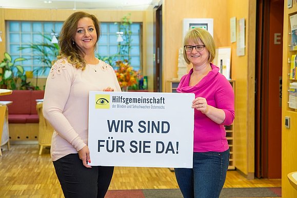 Zwei Mitarbeiterinnen der Hilfsgemeinschaft halten ein Schild mit der Aufschrift "Wir sind für Sie da".