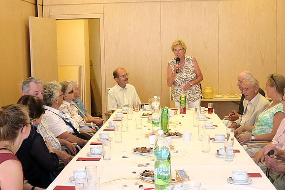 Viele Menschen sitzen an einem Tisch und eine Frau spricht in ein Mikrofon 