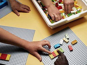 Drei Paar Kinderhände greifen zu bunten Lego Steinen, die auf einem grauen Steckbrett stecken. Die Noppen der Legosteine entsprechen Buchstaben der Braille Schrift. Die jeweiligen Buchstaben sind auch in Schwarz auf den Steinen aufgedruckt. 