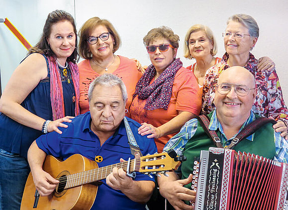 Weibliche Mitglieder der Singgruppe stehen hinter zwei Musikern mit Gitarre und Ziehharmonika