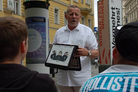 Ein Mann steht in den Straßen von Wien mit einer Mappe in der Hand vor einer Gruppe und spricht.