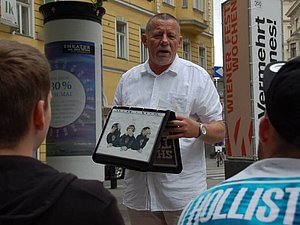Ein Mann steht in den Straßen von Wien mit einer Mappe in der Hand vor einer Gruppe und spricht.