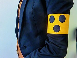 Oberkörper von Person seitlich mit dunkelblauem Jacket und gelber Armbinde mit 3 blauen Punkten