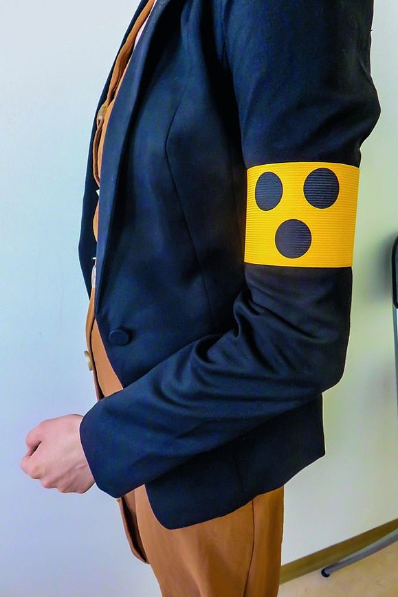 Oberkörper von Person seitlich mit dunkelblauem Jacket und gelber Armbinde mit 3 blauen Punkten
