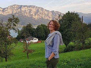 Eine Frau steht auf einem Hügel und blickt auf einem See mit dahinterliegender Bergkette.