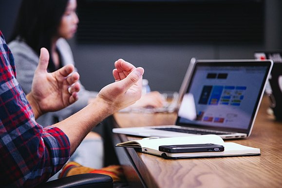 Wir suchen Verstärkung in der Kommunikation! Foto: Unsplash Meetingsituation: gestikulierende Hände und Laptop am Tisch mit Handy