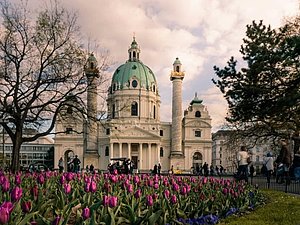 Zentral sieht man die Karlskirche mit ihrem türkisen Kuppeldach und den zwei dünnen Türmen. Im Vordergrund ist ein Blumenbeet mit bunten Tulpen.