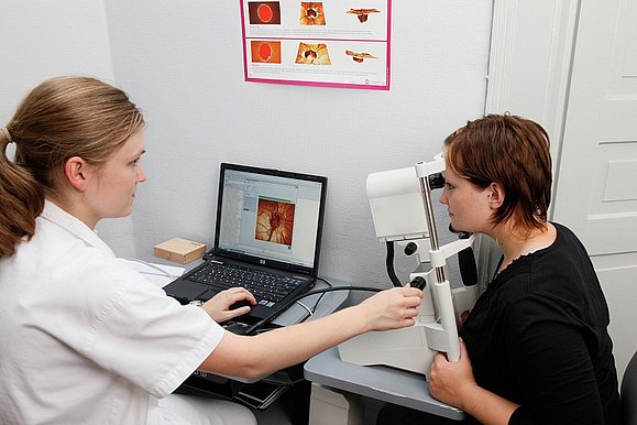 Eine Ärztin führt bei einer rechts von ihr sitzenden Frau eine Augenuntersuchung durch, vor ihr steht ein Computer