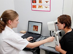 Eine Ärztin führt bei einer rechts von ihr sitzenden Frau eine Augenuntersuchung durch, vor ihr steht ein Computer