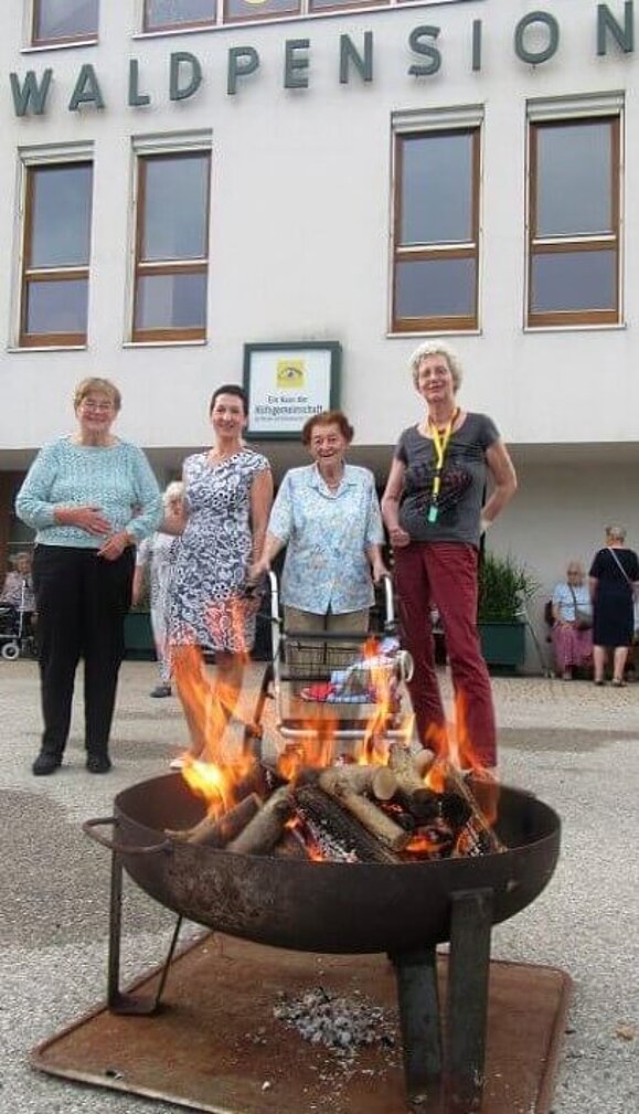 In einer Feuerschale brennt ein großes Feuer, 4 Damen stehen dahinter und schauen zu.