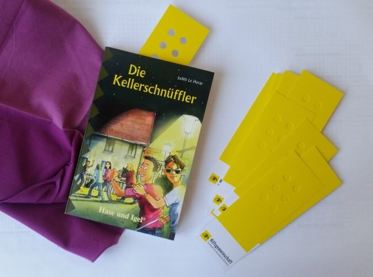 Das Jugendbuch die "Kellerschnüffler" und Lesezeichen mit Braillemuster liegen auf einem Tisch. 