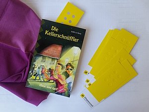 Das Jugendbuch die "Kellerschnüffler" und Lesezeichen mit Braillemuster liegen auf einem Tisch. 